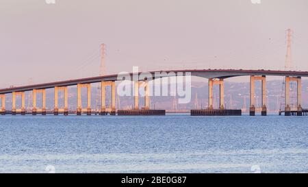 Blick auf den Sonnenuntergang auf die San Mateo Brücke, die die Halbinsel mit der East Bay in der San Francisco Bay Area, Kalifornien verbindet; Stromtürme und Stromleitungen sichtbar Stockfoto