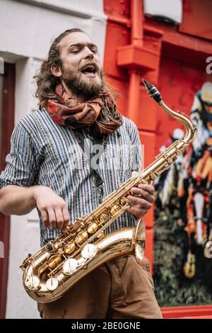 Vertikales Foto eines bärtigen Saxophonisten mit langen blonden Haaren, der in der Mitte der Straße mit emotionalem Ausdruck singt Stockfoto