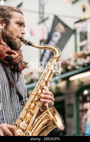 Vertikales Foto eines Saxophonisten bärtig und mit blonden Haaren spielend mit hellen Emotionen auf der Straße voller Menschen Stockfoto