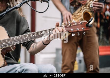 Hand eines jungen Musikers, der Gitarre spielt mitten auf der Straße vor einem Mikrofon und mit einem Saxophonisten im Hintergrund unscharf Stockfoto