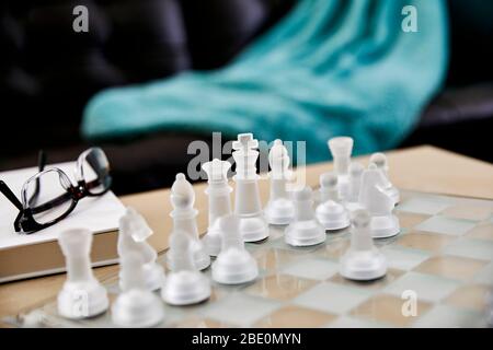 Nahaufnahme von Schachfiguren aus mattiertem Glas auf einem Schachbrett aus Glas in einem Wohnzimmer mit Lesebrille und einem Hardcover-Buch und geringer Schärfentiefe Stockfoto