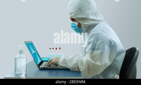 Seitenansicht des Arztes mit blauer Maske und Bioprotective-Anzug, der mit seinem Laptop in seinem Büro arbeitet, mit seinen Händen, die mit Concentrati auf der Tastatur tippen Stockfoto