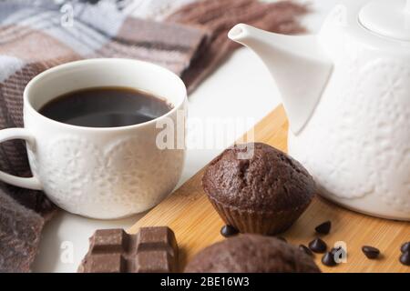 Schokoladenmuffins, Schokoladenchips, Schokoriegel, Tasse Kaffee, Teekanne auf einer Holzfläche mit warmem Schal, Weihnachtsbeleuchtung Bokeh und Stolly in der Stockfoto