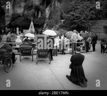 Ein Priester kniet zum Beten. Vor ihm Menschen im Rollstuhl. Im Hintergrund rezitiert ein anderer Priester Gebete am Eingang der Massabielle Stockfoto