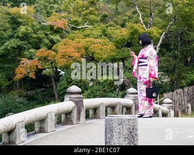 Kyoto, Japan - 30. September 2015: Junge Frau in farbenfrohem Kimono, die ihr Handy überprüft - im Maruyama Park Stockfoto