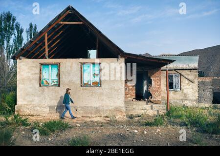 Frau mit Hut und kariertes Hemd zu Fuß in der Nähe von zerstörten alten Haus im Dorf Stockfoto