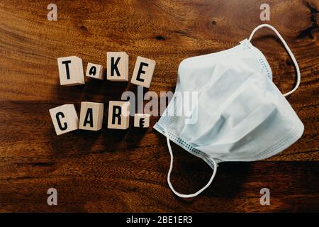 'Take care' Text aus Holz mit medizinischem Maskenwürfel auf Holzhintergrund. Stockfoto