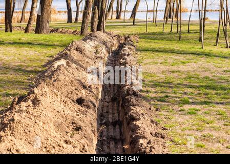 Ein Graben im Boden für die Verlegung eines Rohres oder Rohre, im Park, unter den Bäumen, in der Nähe des Flusses, mit Frühlingslicht Stockfoto