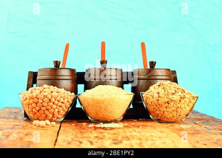 Kichererbsen, Reis und Haferflocken in gläsernen transparenten Tellern auf einem alten Holztisch in kleinen Holzfässern auf türkisfarbenem Hintergrund. Stockfoto