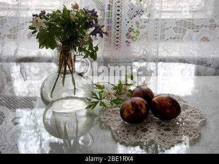 Ein Blumenstrauß aus Frühlingsblumen in einem Glas, bemalte Ostereier an einer gestrickten Serviette vor dem Hintergrund weißer Spitzen-Vorhänge. Stockfoto