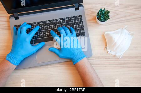 Männliche Hände in blauen Schutzhandschuhen arbeiten auf modernen Laptop. Facharzt in blauen Latexhandschuhen. Medizinische Maske ist auf dem Schreibtisch. Virus und Bacter Stockfoto