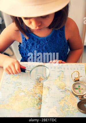 Trendy kleines Mädchen im Sommer Casual Wear und weißen Hut, Blick auf die Weltkarte mit Lupe und Kompass. Planen einer Reise. Entdeckung des Wor Stockfoto
