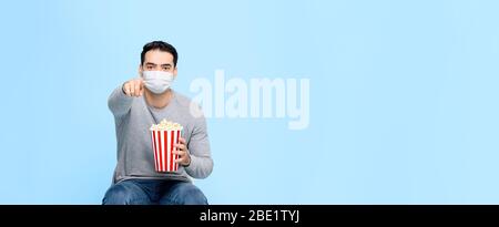 Junger Mann mit Popcorn schützen sich durch das Tragen medizinischer Gesichtsmaske während genießen Film isoliert auf blauem Banner Hintergrund mit Kopierraum Stockfoto