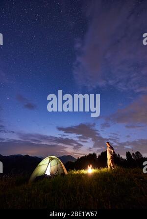 Männliche Touristen verbringen Zeit in den Bergen am Abend mit beleuchteten Zelt und einem Lagerfeuer unter schönen Himmel voller Sterne. Nachtcamping in den Bergen im Sommer Stockfoto