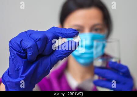 Eine Frauenhand in einer Schutzmaske und ein blauer medizinischer Handschuh hält mit ihren Fingern eine weiße Tablette. Nahaufnahme Stockfoto