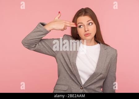 Tötet mich bitte. Porträt einer jungen Frau im Geschäftsanzug erschöpft von der Arbeit, Zeigen mit der Fingerpistole auf den Kopf, zeigen Selbstmord Geste, schießen sich selbst. Stockfoto