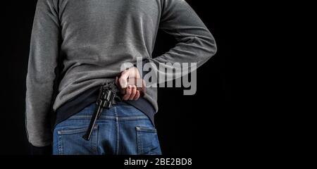 Nahaufnahme eines Mannes, der eine Waffe hinter seinem Rücken hält. Ein Mann in Jeans und ein graues Sweatshirt mit Waffen auf dunklem Hintergrund. Stockfoto