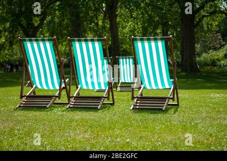 Eine Gruppe von bunt gestreiften traditionellen Klappliegen, die leer in einem sonnigen grünen Graspark in London, Großbritannien, sitzen Stockfoto