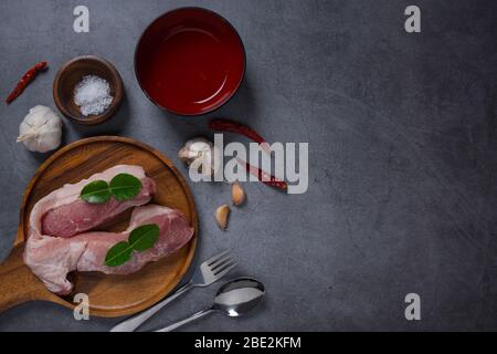 Frisches Fleisch, Rind oder Schweinefleisch auf Holzblock mit Zutaten zum Kochen auf dunklem Zementgrund Stockfoto