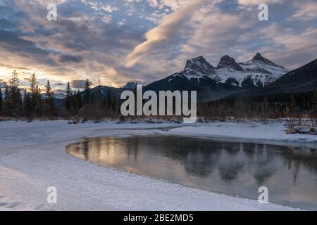 Schneebedeckte Gipfel der Three Sisters spiegeln sich in ruhigem, eisigen Wasser, Morgenstimmung, Bow River, Canmore, Banff National Park, Alberta, Kanada Stockfoto