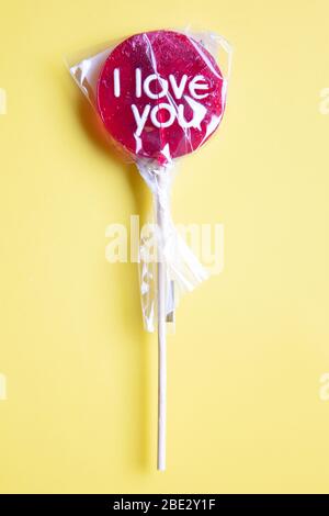 Ein süßes gewickeltes 'I Love You' rotes Lollypop vor pastellgelbem Hintergrund, flaches Lay, Draufsicht, minimalistisch Stockfoto
