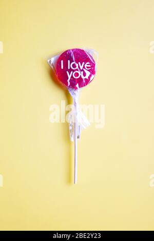 Ein süßes gewickeltes 'I Love You' rotes Lollypop vor pastellgelbem Hintergrund, flaches Lay, Draufsicht, minimalistisch Stockfoto