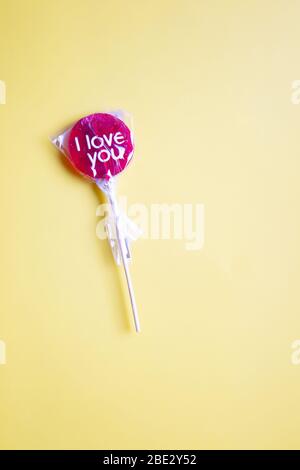 Ein süßes gewickeltes 'I Love You' rotes Lollypop vor pastellgelbem Hintergrund, flaches Lay, Draufsicht, minimalistisch Stockfoto