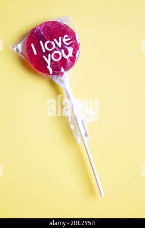 Ein süßes gewickeltes 'I Love You' rotes Lollypop vor pastellgelbem Hintergrund, flaches Lay, Draufsicht, minimalistisch Stockfoto