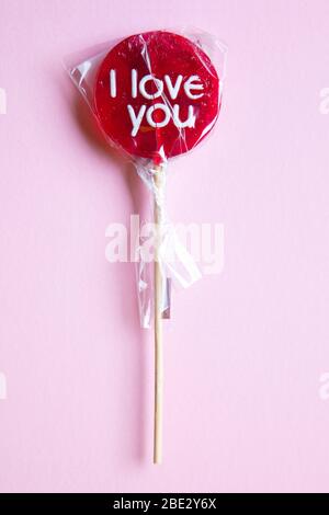 Ein süßes gewickeltes 'I Love You' rotes Lollypop vor einem pastellrosa Hintergrund, flaches Lay, Draufsicht, minimalistisch Stockfoto