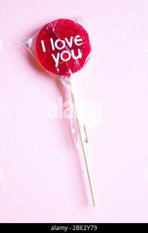 Ein süßes gewickeltes 'I Love You' rotes Lollypop vor einem pastellrosa Hintergrund, flaches Lay, Draufsicht, minimalistisch Stockfoto