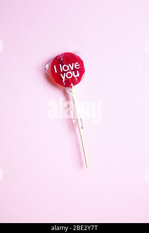 Ein süßes gewickeltes 'I Love You' rotes Lollypop vor einem pastellrosa Hintergrund, flaches Lay, Draufsicht, minimalistisch Stockfoto