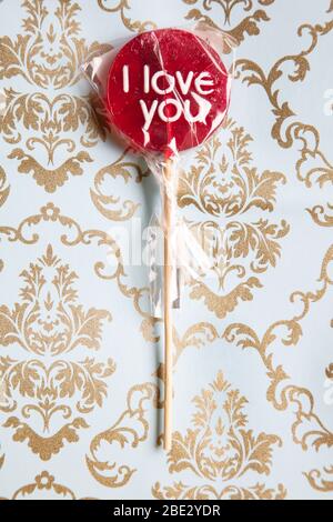 Ein süßes gewickeltes 'I Love You' rotes Lollypop vor einem Gold und hellblau dekorierten Musterhintergrund, flaches Lay, Draufsicht, minimalistisch Stockfoto