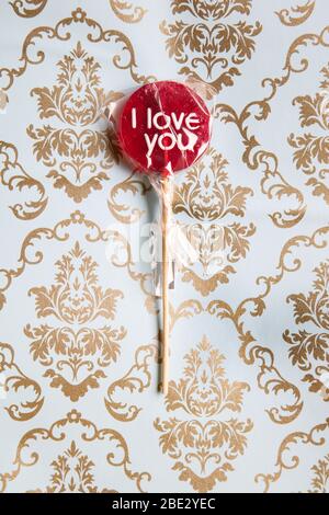 Ein süßes gewickeltes 'I Love You' rotes Lollypop vor einem Gold und hellblau dekorierten Hintergrund, flaches Lay, Draufsicht, minimalistisch Stockfoto