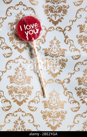 Ein süßes gewickeltes 'I Love You' rotes Lollypop vor einem Gold und hellblau dekorierten Hintergrund, flaches Lay, Draufsicht, minimalistisch Stockfoto