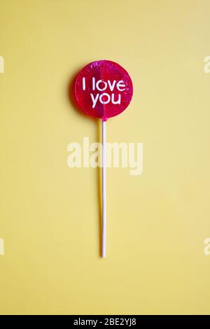 Ein ungewickelter 'I Love You' roter Lollypop vor pastellgelbem Hintergrund, flaches Lay, Draufsicht, vertikal Stockfoto