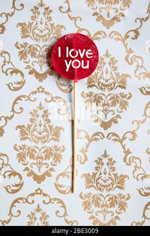 Ein süßes 'I Love You' rotes Lollypop vor einem Gold und hellblau dekorierten Damasthintergrund, flaches Lay, Draufsicht, minimalistisch, vertikal Stockfoto