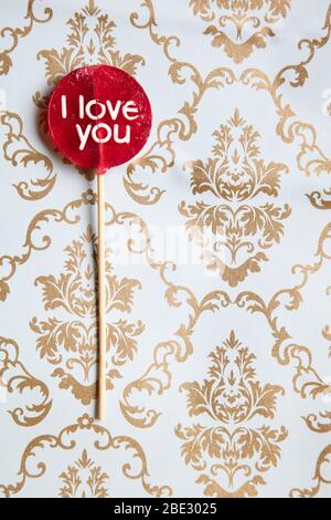 Ein süßes 'I Love You' rotes Lollypop vor einem Gold und hellblau dekorierten Hintergrund, flaches Lay, Draufsicht, minimalistisch, vertikal Stockfoto