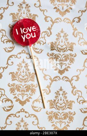 Ein süßes 'I Love You' rotes Lollypop vor einem Gold und hellblau dekorierten Hintergrund, flaches Lay, Draufsicht, minimalistisch, vertikal Stockfoto