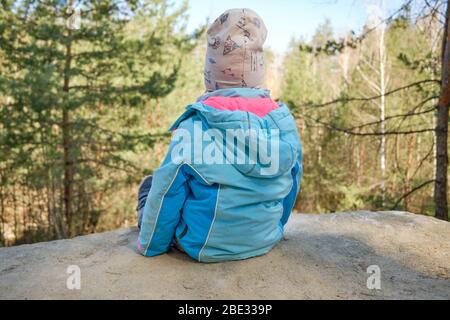 Rückansicht des 4-jährigen kaukasischen Kindes, das auf Sandsteinfelsen sitzt und an einem schönen Frühlingstag in Deutschland den Blick auf den Wald genießt Stockfoto