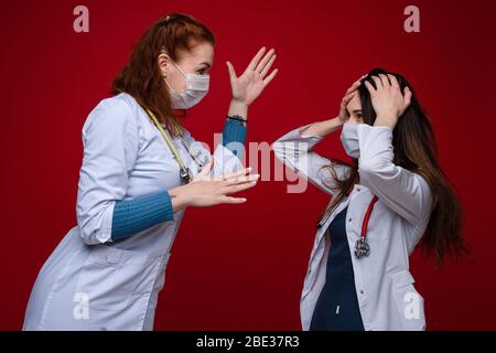 Porträt von zwei Ärztinnen in medizinischer Kleidung mit Phonendoskop um den Hals streiten, Bild isoliert auf rotem Hintergrund Stockfoto
