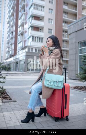 Dunkelhaarige junge Frau in einem beigefarbenen Mantel sitzt auf ihrem Koffer Stockfoto