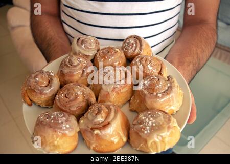 Ein Mann hält hausgemachte frisch gebackene Cinnabon Buns mit Sahne-Sauce und Zimtpulver auf einem großen weißen Teller auf hellem unscharfen Hintergrund. Hausgemachte Bäckerei. Stockfoto