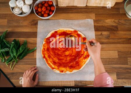 Draufsicht auf die Hände einer Frau, die Tomatensauce zu einem Pizzateig auf einer Holzarbeitspfläche hinzufügt. Roher Teig für die Pizzazubereitung mit Zutat: Tomatensauce, m Stockfoto