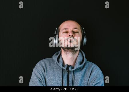 Bärtiger entspannter Mann im Hoodie, der mit Kopfhörern vor schwarzem Hintergrund steht, Musik hört, Kopf und Schultern Stockfoto