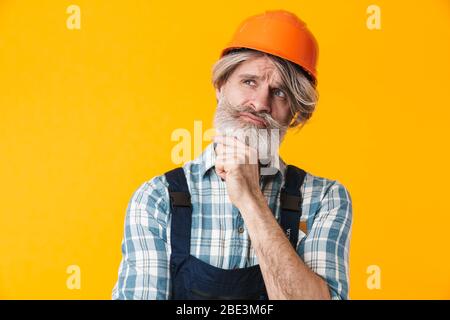Foto von ernsthaften Denken ältere grauhaarige bärtige Mann Baumeister in Helm posiert isoliert über gelben Wand Hintergrund. Stockfoto