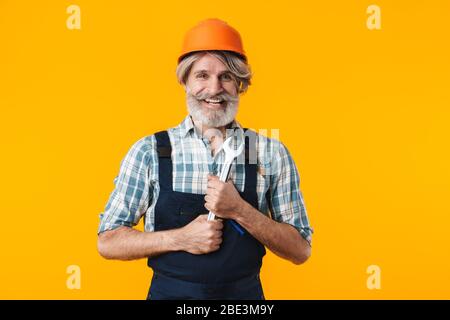 Bild von glücklich älteren grauhaarigen bärtigen Mann Baumeister im Helm posiert isoliert über gelbe Wand Hintergrund halten Schraubenschlüssel. Stockfoto