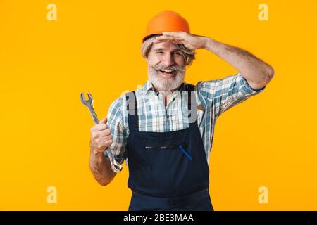 Bild von glücklich älteren grauhaarigen bärtigen Mann Baumeister im Helm posiert isoliert über gelbe Wand Hintergrund halten Schraubenschlüssel. Stockfoto