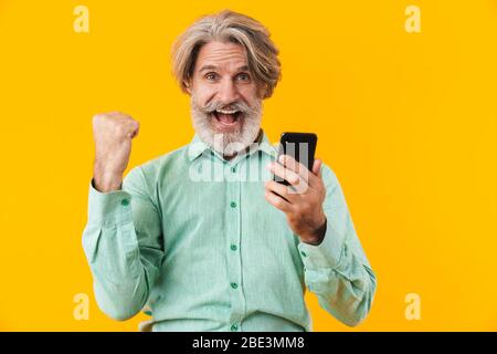 Foto von fröhlichen überrascht grauhaarigen bärtigen Mann in blauem Hemd posiert isoliert über gelbe Wand Hintergrund mit Handy machen Gewinner Geste. Stockfoto
