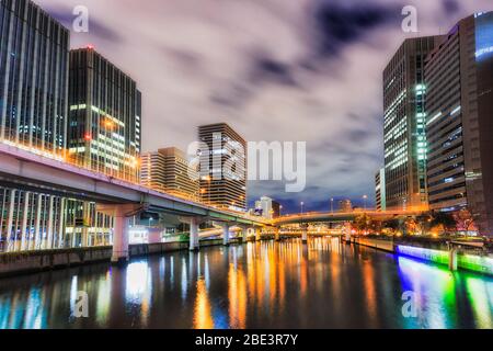 Dunkle Fluss- und Autobahnbrücken über der Wasseroberfläche zwischen Hochhäusern in Osaka bei Sonnenaufgang. Stockfoto