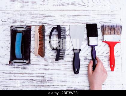 Alte Werkzeuge (Pinsel, Spachtel, Drahtbürste, Sandpapierhalter) und Hand, die einen brus One auf weißem Rand nimmt. Blick von oben, Stockfoto